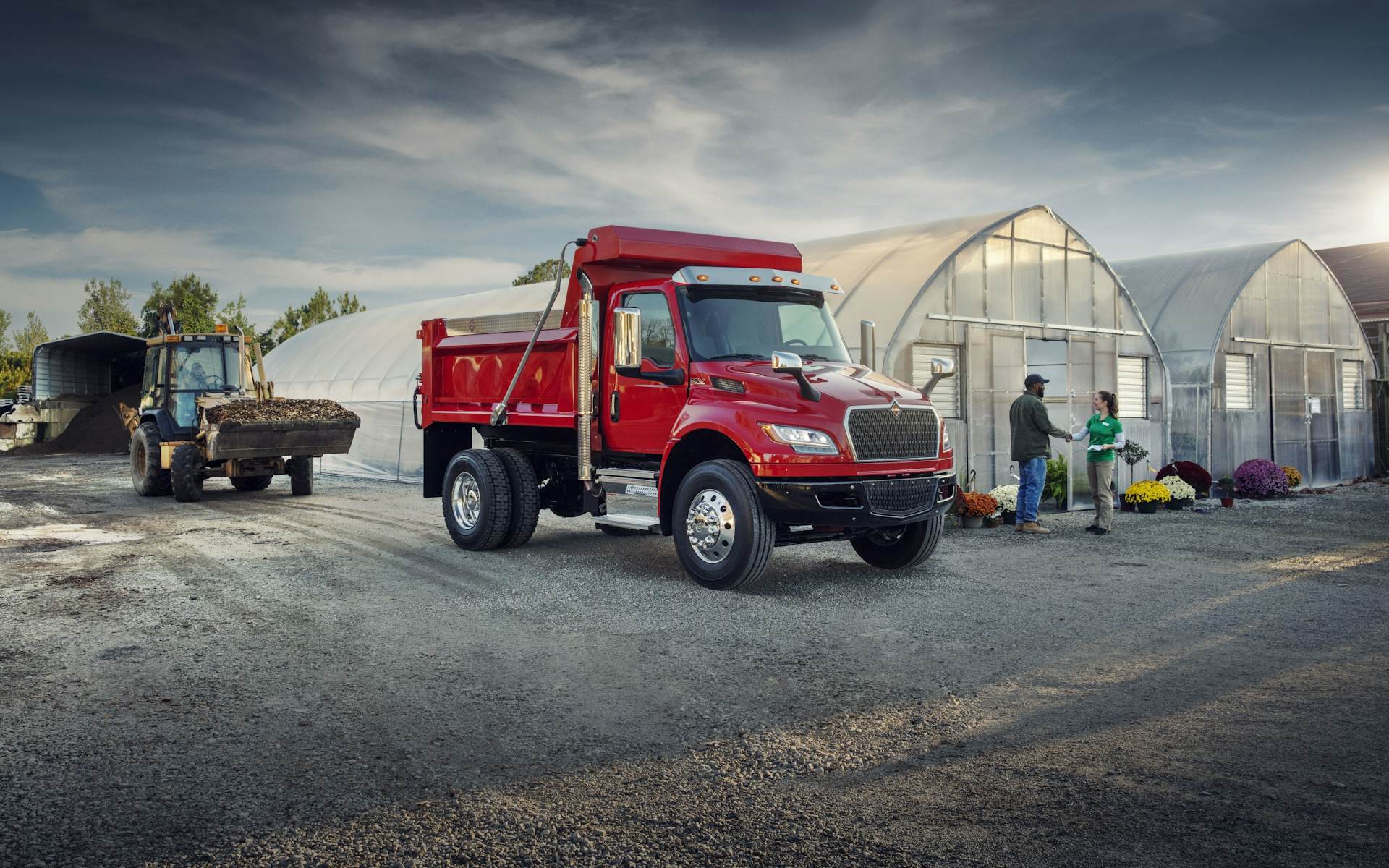 MV dump truck parked on gravel next to a greenhouse and two people