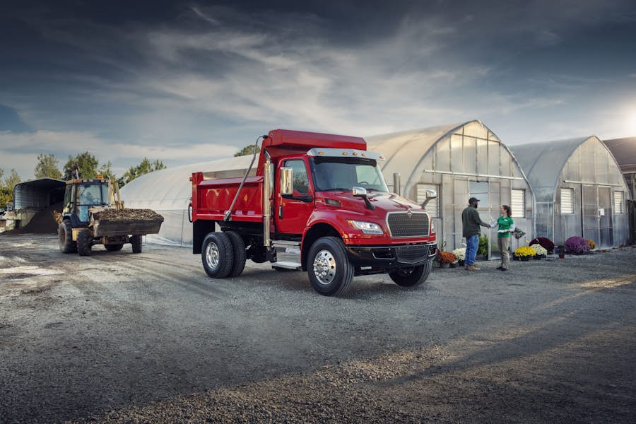 MV dump truck parked on gravel next to a greenhouse and two people